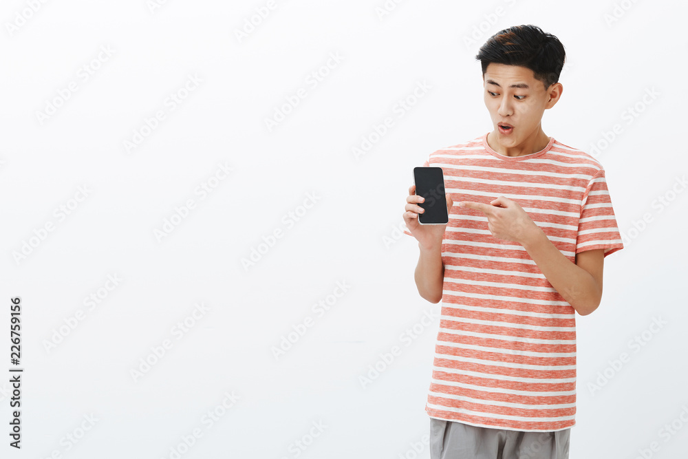 Portrait of impressed and surprised excited young attractive chinese guy in striped t-shirt open mouth from interest and thrill holding smartphone pointing and looking at cellphone screen fascinated Portrait of impressed and surprised excited young attractive chinese guy in striped t-shirt open mouth from interest and thrill holding smartphone pointing and looking at cellphone screen fascinated