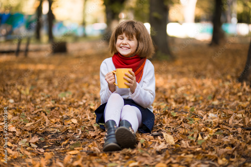 Happy little girl drinks hot tea in autumn park in surround of yellow