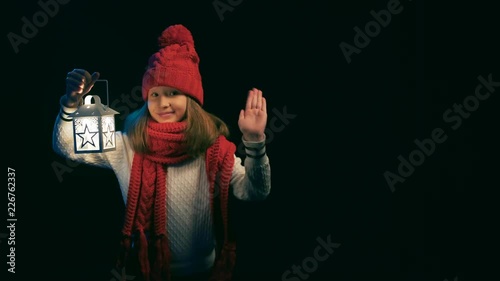 Little girl wearing knitted winter hat and scarf holding a lantern looking at camera smiling and waving a hand than walikng away, over black background photo