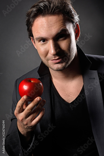 Handsome and cool man with mysterious look sholding an apple while smiling. Hot and sexy guy in a dark jacket with a confident face. © Антон Зимин