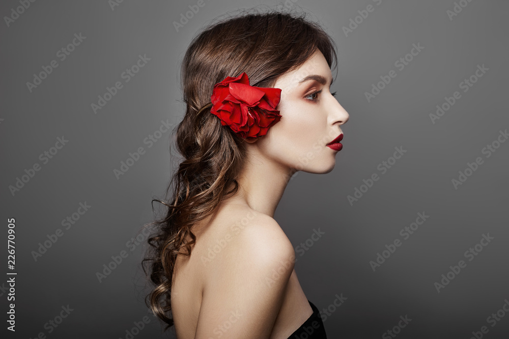 Woman with a big red flower in her hair. Brown-haired girl with a red flower posing on a gray background. Big beautiful eyes and natural makeup. Long curly hair, perfect face Woman with a big red flower in her hair. Brown-haired girl with a red flower posing on a gray background. Big beautiful eyes and natural makeup. Long curly hair, perfect face