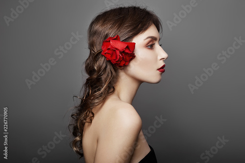 Woman with a big red flower in her hair. Brown-haired girl with a red flower posing on a gray background. Big beautiful eyes and natural makeup. Long curly hair, perfect face