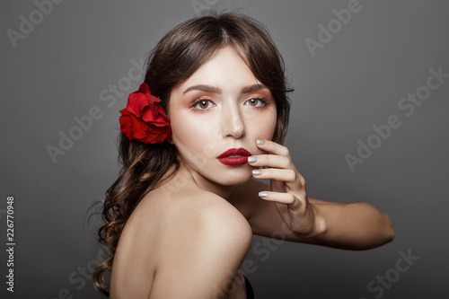 Woman with a big red flower in her hair. Brown-haired girl with a red flower posing on a gray background. Big beautiful eyes and natural makeup. Long curly hair, perfect face