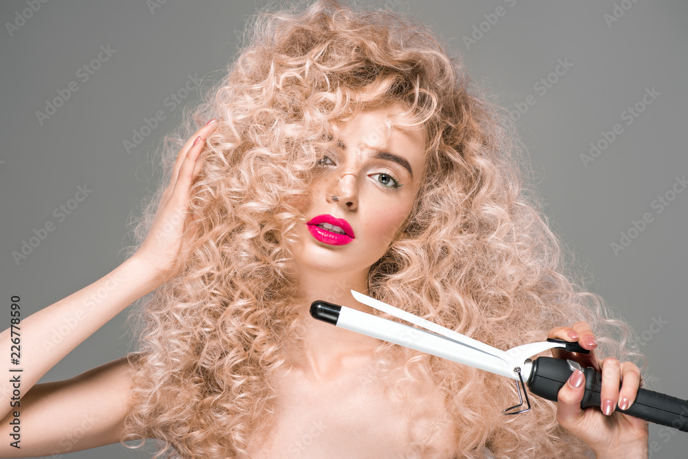 Obraz premium attractive naked girl with long curly hair holding hair curler and looking at camera isolated on grey