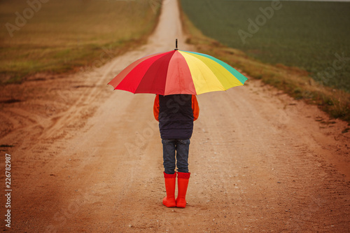 Child in orange rubber boots holding colorful umbrella under rain in autumn. Back view photo