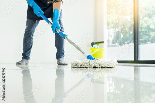 Young housekeeper cleaning floor mobbing holding mop and plastic bucket with brushes, gloves and detergents in the leaving room house floor helping his wife.