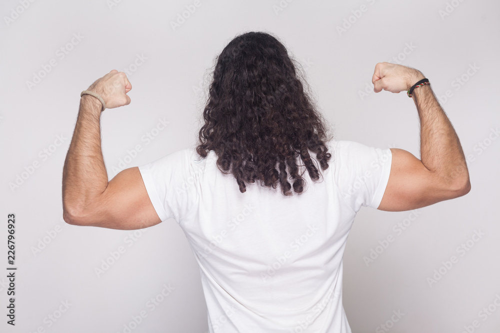 Back side of strong bodybuilder man in white t-shirt with long wavy hair,  standing and