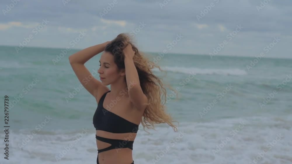 Portrait of a charming young woman in sexy bikini posing on beach, windy day on beach with high waves. Curly hairs floats on a wind, fit girl. Portrait of a charming young woman in sexy bikini posing on beach, windy day on beach with high waves. Curly hairs floats on a wind, fit girl.