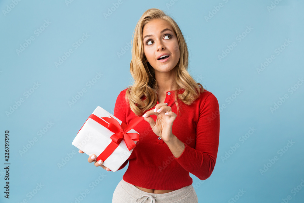 Portrait of happy blond woman 20s wearing red shirt holding present box, isolated over blue background in studio Portrait of happy blond woman 20s wearing red shirt holding present box, isolated over blue background in studio