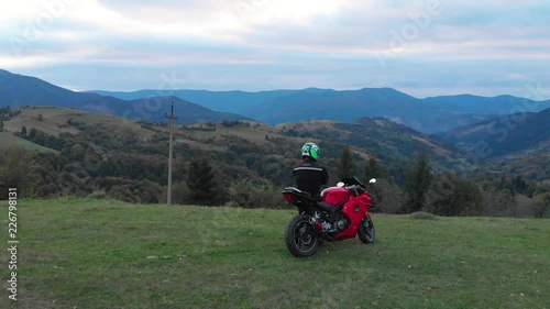 A guy on a motorcycle. Young handsome guy rides a motorcycle on a mountain road. The guy stands near his motorcycle and looks at the mountains. He is happy. Shooting from the quadcopter.