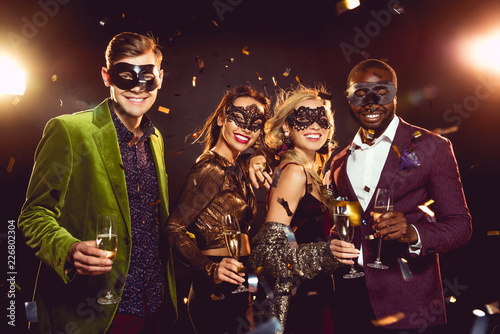 glamorous multiethnic friends in carnival masks holding champagne glasses and celebrating new year on party