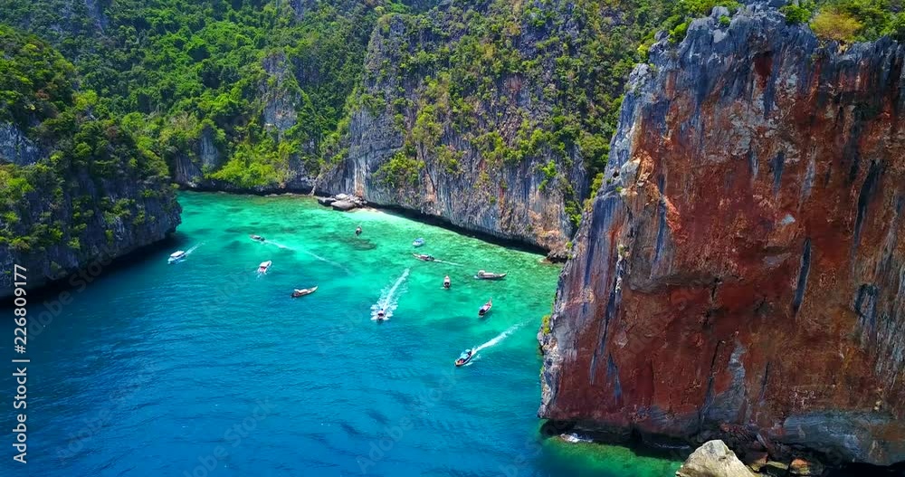 Multiple Boats in Loh Samah Bay, Thailand - Tropical Cove Surrounded by Lush Island Cliff Face With Turquoise Blue Water and Visible Coastal Reef - Traversing Overhead View Multiple Boats in Loh Samah Bay, Thailand - Tropical Cove Surrounded by Lush Island Cliff Face With Turquoise Blue Water and Visible Coastal Reef - Traversing Overhead View