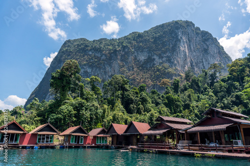 Wooden Thai traditional floating houses on a lake with mountains and rain forest in the background during a sunny day at Ratchaprapha Dam at Khao Sok National Park, Thailand