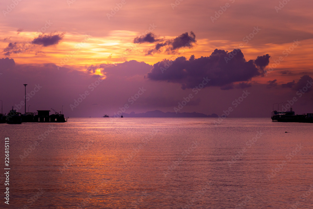 Tropical orange evening sunset and pier. Colorful sky in the dusk. Calm sea and cottage silhouettes above the water.