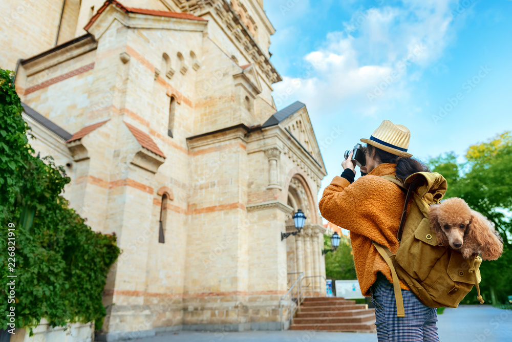 Woman traveler with dog in the backpack taking pictures with a camera while traveling.	