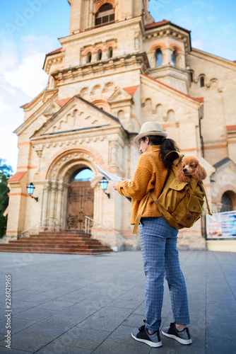 Woman traveler with dog in the backpack examines architectural monument . Concept of travel.