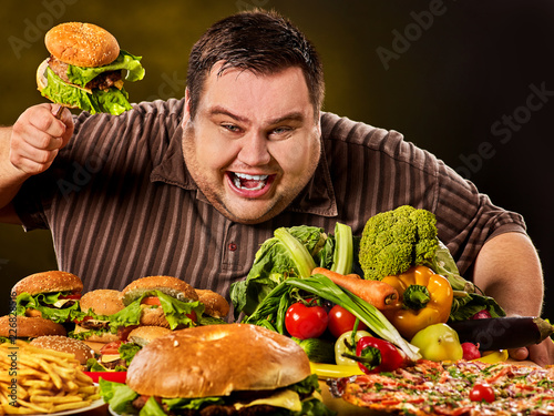 Diet fat man who makes choice between healthy and unhealthy food. Overweight male with hamburgers, french fries and vegetables on black background. Dangers of extra weight
