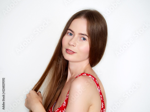 Natural light portrait of young beautiful blond woman with long hair in vintage red dress smiling looking at camera over white wall background.