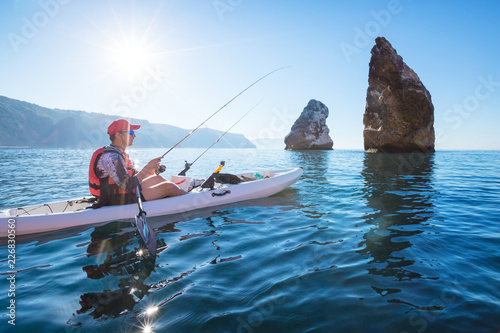 A man fishing on a kayak boat in the sea near the rocks at the shore of island mountain. Young fisherman kayaking.