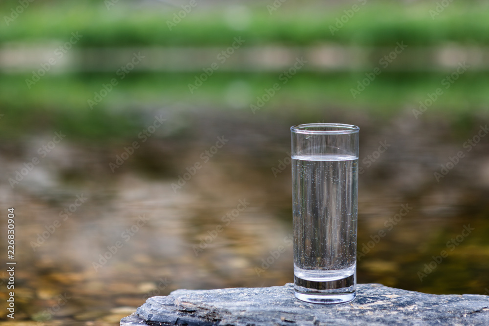 Glass of Cold And Clean Water On Background River And Grass.