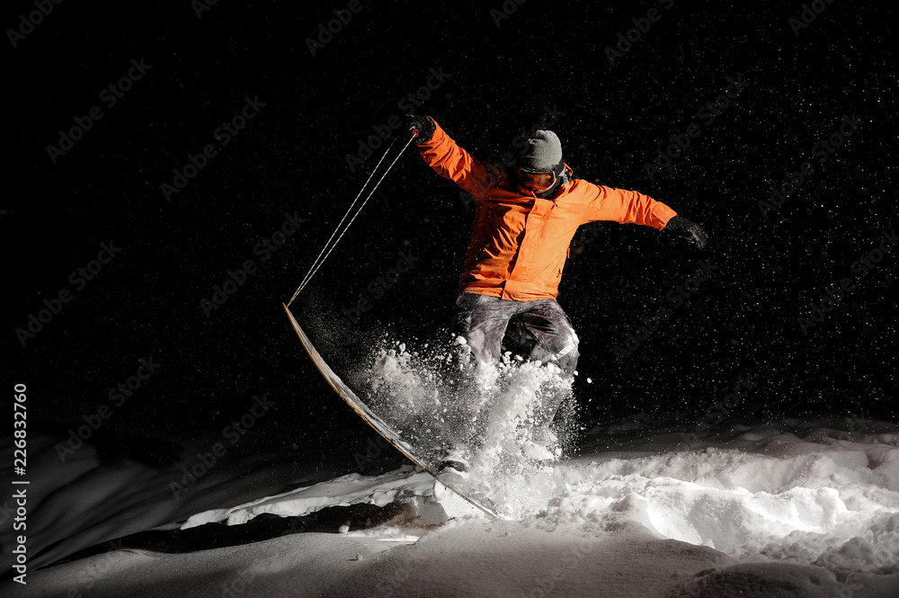 Fototapeta premium Aktywny snowboardzista w pomarańczowym stroju sportowym i masce skaczący nocą po zaśnieżonym wzgórzu