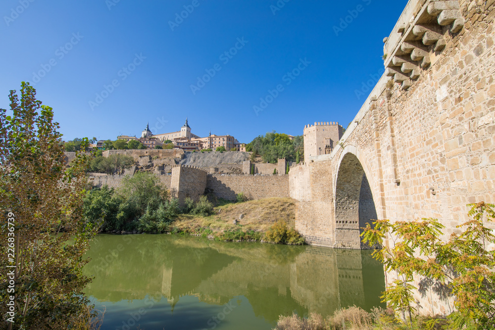 Alcazar from green water river Tagus, Tajo in Spanish, Alcantara arch bridge,  landmark and monument from ancient Roman age, in Toledo city, Spain, Europe