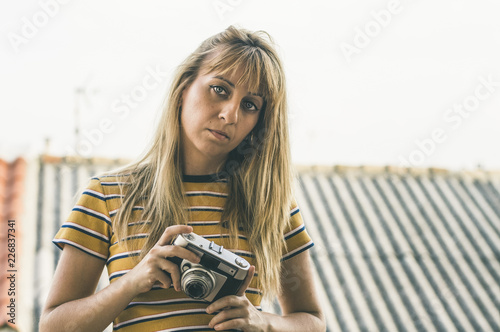 Preciosa chica rubia con pelo largo y ojos azules de frente de medio cuerpo mirando a la cámara sujetando una cámara analógica con camiseta amarilla con rayas  photo