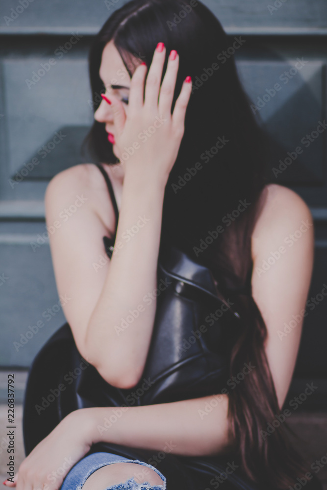 Portrait young beautiful sexy slender girl with dark long straight hair posing in black jacket. Fashion leather clothes hipster concept. Girl sits with a passionate gaze on the street. Portrait young beautiful sexy slender girl with dark long straight hair posing in black jacket. Fashion leather clothes hipster concept. Girl sits with a passionate gaze on the street.