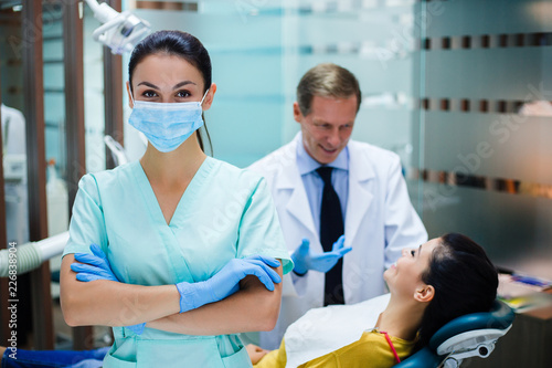 She can help you. Beautiful young nurse in mask looking at camera while standing in dentist’s office