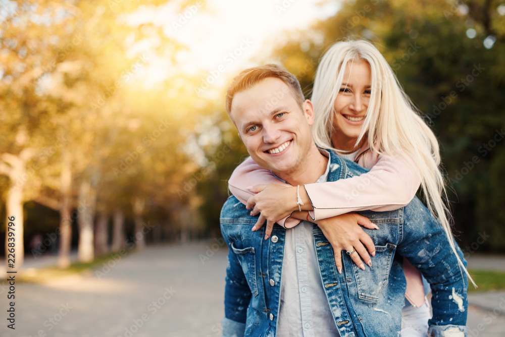 Fototapeta premium Closeup shot of young man carrying young woman on his back. Happy smiling couple looking at camera. Happy couple putdoor having fun piggyback in love.