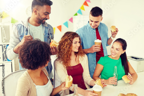 corporate, celebration and people concept - happy friends or team eating sandwiches with coffee and non-alcoholic drinks at office party