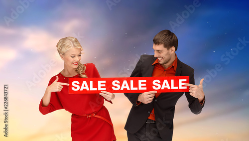 shopping and promotion concept - smiling couple with red sale sign over sky background
