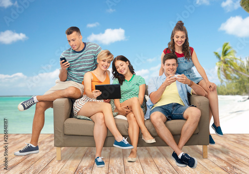 friendship, leisure and technology concept - group of happy smiling friends with tablet pc computer and smartphones sitting on sofa over tropical beach background in french polynesia