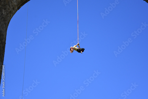 Bungee jumping from railway bridge. Seen from the ground beautiful young woman hanging on a cord high in the blue sky