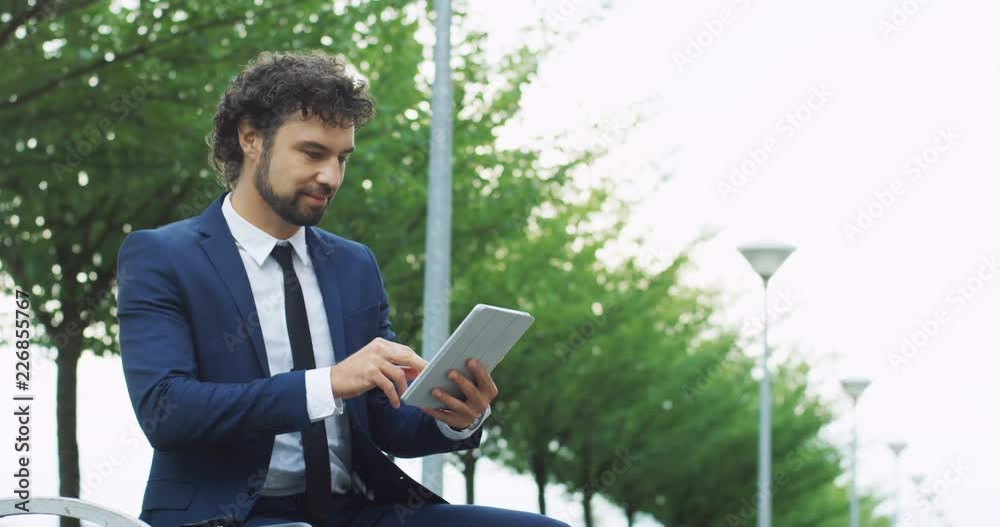 Handsome Caucasian businessman in suit and tie using his tablet device while sitting on the bench outdoor. Handsome Caucasian businessman in suit and tie using his tablet device while sitting on the bench outdoor.
