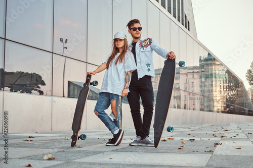 Valokuva Attractive young couple dressed in trendy clothes posing with skateboards near skyscraper