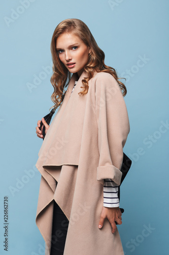 Young attractive woman with wavy hair in beige coat with black backpack thoughtfully looking in camera over blue background isolated