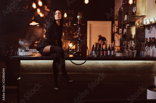 Seductive brunette girl in black clothes sitting with crossed legs on counter in a nightclub.