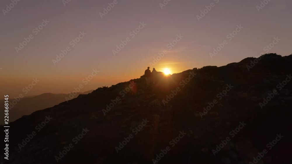 Slow motion aerial drone shot of camera following couple of travellers or hikers on their way to mountain top or peak during amazing beautiful orange sunset or dawn. Motivationaland inspiring moment Slow motion aerial drone shot of camera following couple of travellers or hikers on their way to mountain top or peak during amazing beautiful orange sunset or dawn. Motivationaland inspiring moment