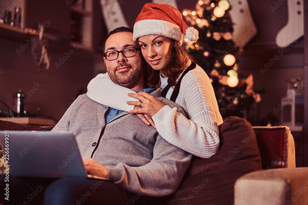 Portrait of an adorable couple with laptop - charming woman in Santa hat hugging her man and looking at camera. Portrait of an adorable couple with laptop - charming woman in Santa hat hugging her man and looking at camera.