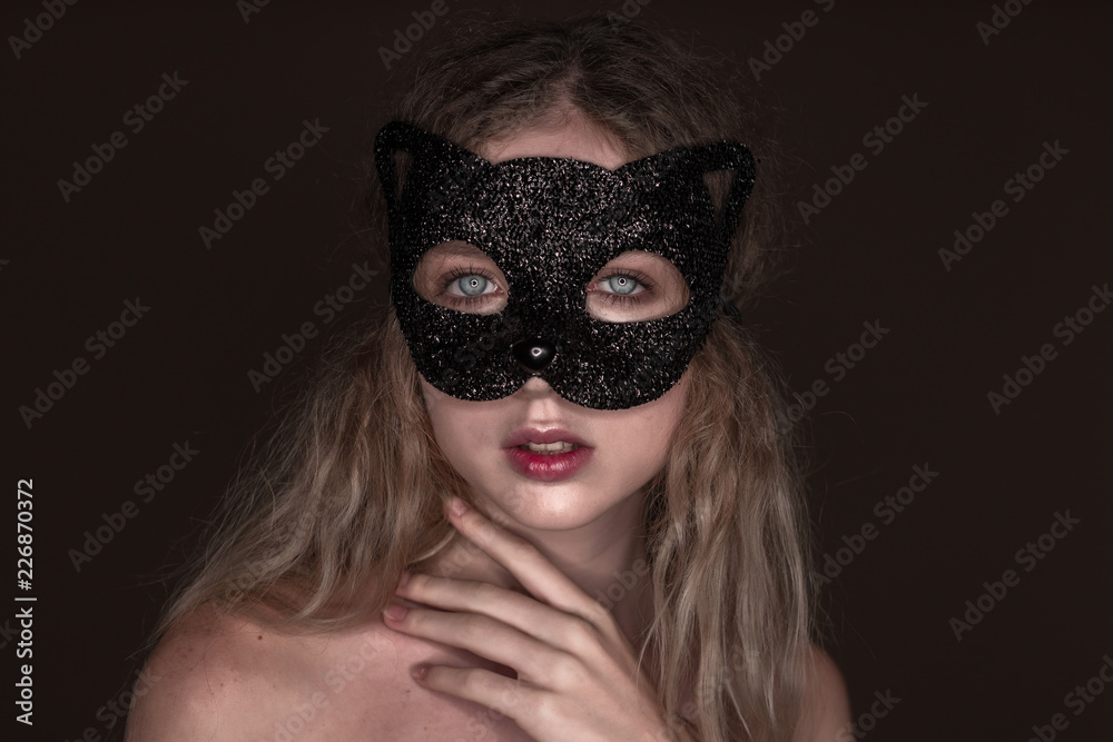 Beautiful young tender woman posing isolated over dark beige chocolate brown background wall wearing carnival cat masquerade mask looking camera.