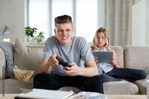 Siblings at home, boy playing video game, girl watching tablet