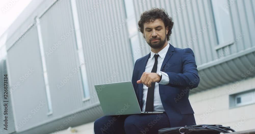 Attractive Caucasian businessman sitting on the bench outside with laptop computer, looking at his watch and going away. Attractive Caucasian businessman sitting on the bench outside with laptop computer, looking at his watch and going away.