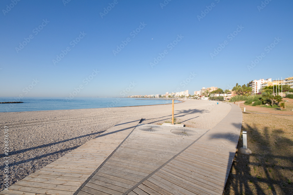landscape Els Terrers Beach, in Benicassim, Castellon, Valencia, Spain, Europe. Palm trees in shadows, wooden boardwalk, buildings, blue clear sky and Mediterranean Sea