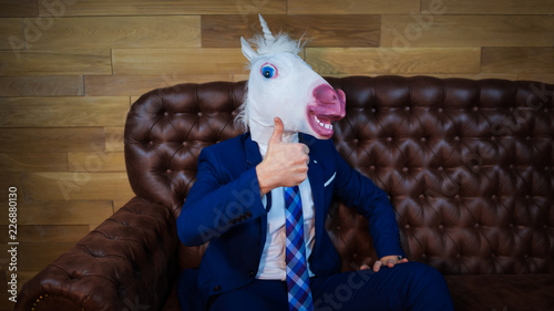 Funny unicorn in elegant suit sits on sofa like a boss and showing gesture thumbs up. Portrait of unusual man at home. Freaky young manager in comical mask.