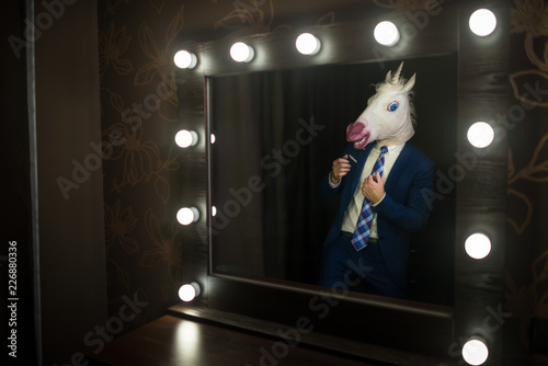 Glamour guy in elegant suit and comical mask looks at himself in the mirror in dressing room. Freaky young man in stylish room posing like a boss. Unusual unicorn.
