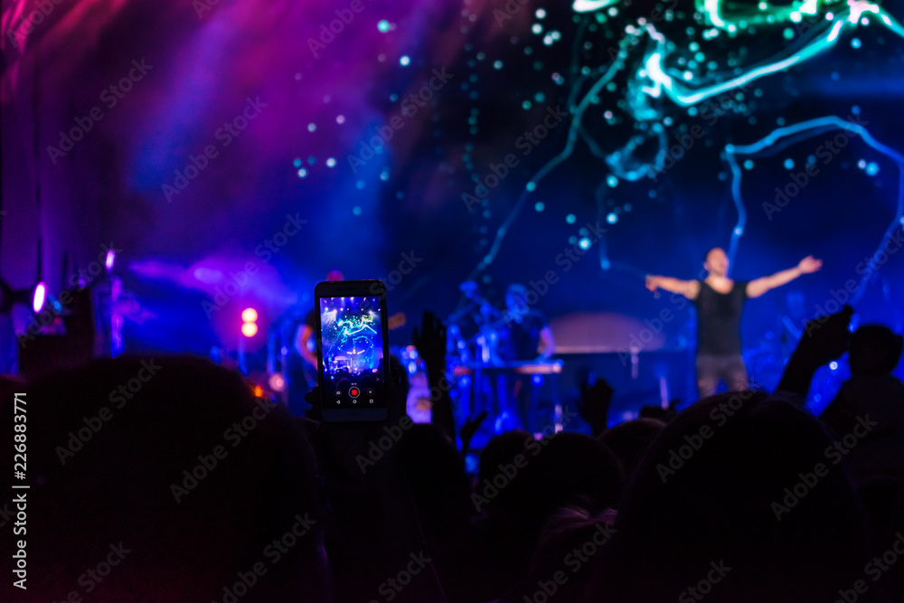 Crowd at concert. People silhouettes on backlit by bright blue and purple stage lights. Cheering crowd in colorful stage lights. Raised hands and smartphones against scene