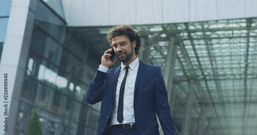 Caucasian young attractive man in the suit and tie standing outside big urban building and answering his moile phone ringing with a smile. Caucasian young attractive man in the suit and tie standing outside big urban building and answering his moile phone ringing with a smile.