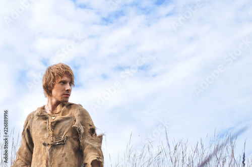 young man dressed as a medieval peasant amidst wild yellowed ears against a blue sky