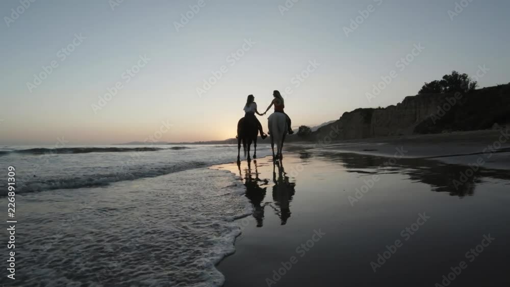 Tracking shot, women hold hands horseback riding on beach